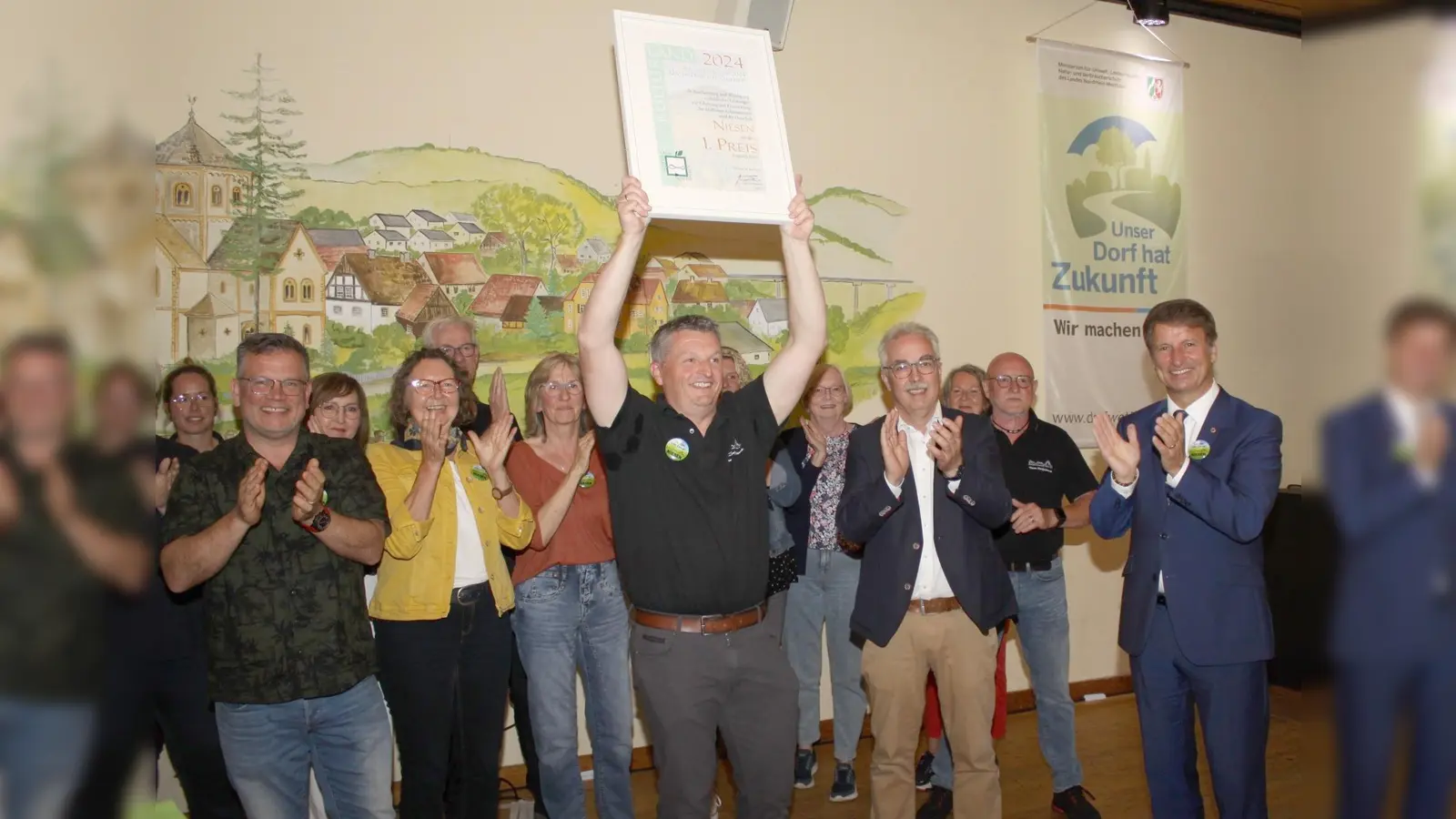 Markus Hagemann, Ortsbürgermeister von Niesen, präsentiert stolz die Siegerurkunde. Dafür gab es großen Applaus von Landrat Michael Stickeln (rechts), Willebadessens Bürgermeister Norbert Hofnagel (Zweiter von rechts), den glücklichen Bürgerinnen und Bürgern Niesens und den zahlreichen Gästen in der Bürgerhalle. (Foto: Kreis Höxter)